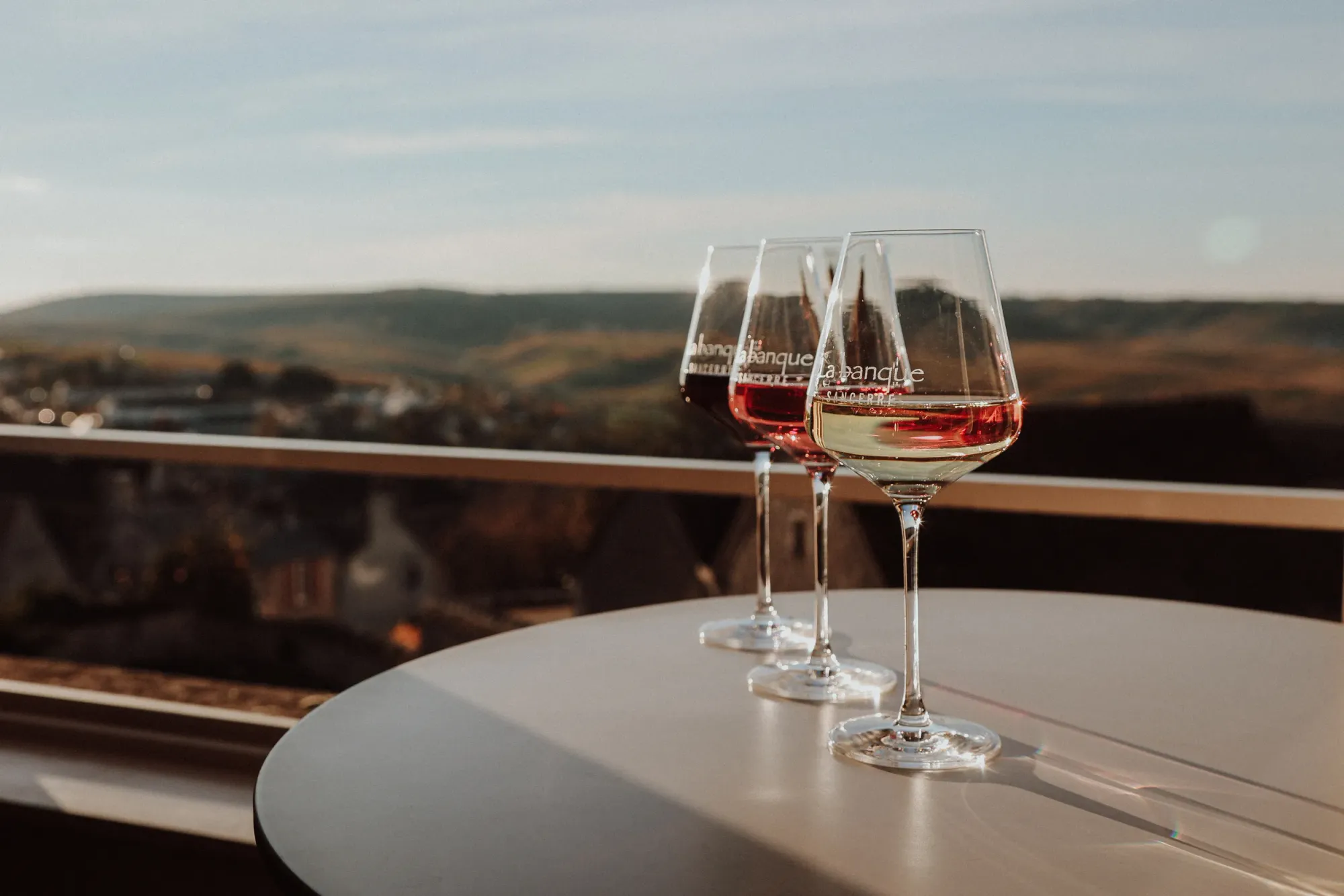 Trois verres gravés La Banque, rouge, rosé et blanc, sur une table en terrasse face aux collines du Sancerrois